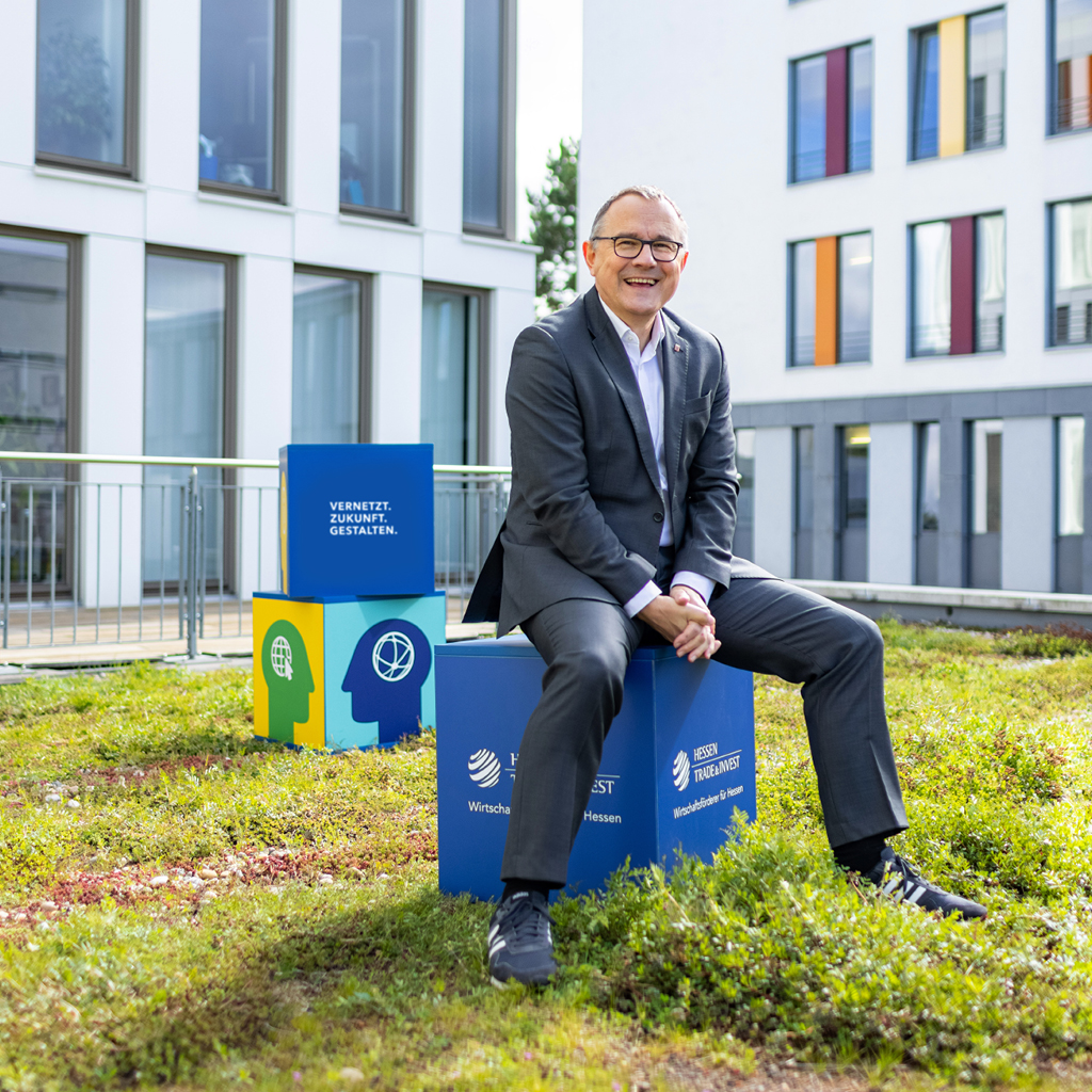 Dr. Rainer Waldschmidt sitzt locker auf einem blauen Würfel mit dem Logo von Hessen Trade & Invest und lächelt in die Kamera. Der Würfel steht auf einem Rasenstück, weiter hinten stehen zwei farbige Würfel übereinander. Auf dem unteren sind illustrierte Köpfe im Profil zu sehen. Den Hintergrund bildet ein Gebäude mit vielen schmalen hohen Fenstern.