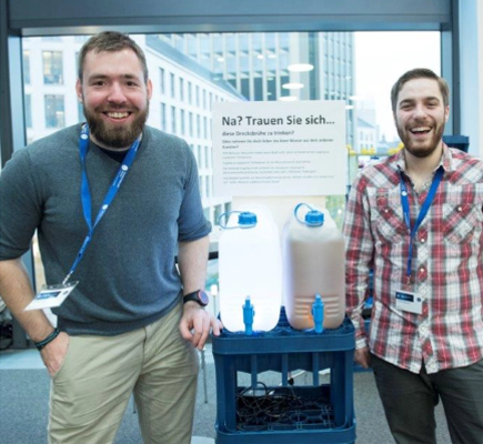 Zwei lächelnde Studenten vor zwei gefüllten Wasserkanistern aus Plastik. Der linke Kanister enthält sauberes, klares Wasser, der rechte schmutziges braunes Wasser.